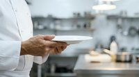 Chef in white uniform holding empty plate in professional kitchen environment with blurred background, suitable for culinary, restaurant, hospitality and food service concepts.