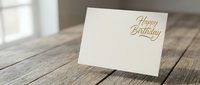 Elegant cream birthday greeting card with gold lettering standing on rustic wooden table surface in soft natural lighting for celebration designs.