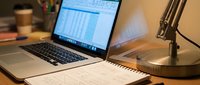 Professional workspace with laptop displaying spreadsheet data, notebook, desk lamp and coffee cup for business productivity and financial analysis.