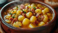 Small round potatoes with bacon pieces in creamy sauce, garnished with herbs and black pepper, served in rustic ceramic bowl. Traditional comfort food.