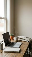 Minimalist workspace with open laptop, ceramic mug, succulent plant, and folded blanket on wooden desk by bright window with neutral beige wall for home office or creative work.