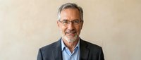 Mature male professional with gray hair and glasses wearing dark blazer over light blue shirt, smiling at camera against neutral beige background for corporate portraits.