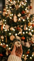 Closeup of hand holding festive ornament with decorated evergreen tree and warm lights blurred in background, perfect for holiday greeting cards and seasonal marketing.