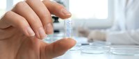 Laboratory scientist in white coat examining small glass vial with clear liquid sample in modern research facility for pharmaceutical development and testing.