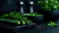 Fresh green cucumbers and dill on black wooden cutting board with scattered peppercorns and herbs in dark moody setting for food photography.