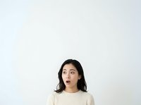 Young woman with dark shoulder-length hair and East Asian ethnicity wearing a cream ribbed sweater, looking upward with surprised expression against neutral white background, isolated for design use.