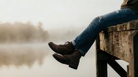 Person in jeans and boots sitting on wooden dock by calm lake during golden hour sunrise creating peaceful morning atmosphere for relaxation concepts.