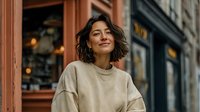 Young woman with short brown hair wearing beige sweater smiling outdoors on city street with storefronts and warm lighting in urban setting.