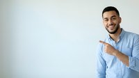 Young Middle Eastern man with dark hair and beard wearing light blue button-up shirt, smiling and pointing to blank white space with both hands for advertising or promotional content.