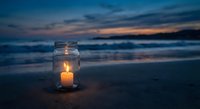 Glowing candle in glass jar on sandy beach at twilight with ocean waves and colorful sunset sky creating peaceful romantic atmosphere for meditation.