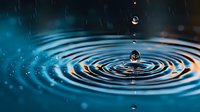 Water drop falling into calm surface creating perfect concentric ripples with orange and blue reflections in macro photography. Frozen motion captures multiple droplets.
