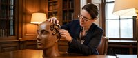 Caucasian female scientist adjusting golden gears inside a wooden human head sculpture in a classic library, symbolizing mind and intelligence.
