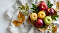 Fresh red, yellow, and green apples in wooden bowl with autumn leaves on white linen tablecloth, creating rustic harvest scene.