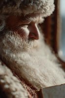 Elderly Caucasian man with white beard in warm winter hat looking thoughtfully out window during cold season reflecting on memories and life experiences.
