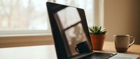 Modern laptop computer with blank black screen on wooden desk beside small potted succulent plant and coffee mug in bright home office workspace.