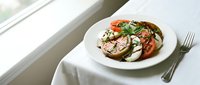 Fresh caprese salad with ripe tomatoes, mozzarella, and herbs on a white plate, served on a linen tablecloth near a bright window.