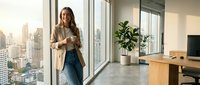 Young professional businesswoman standing by office window with city skyline view, holding smartphone in modern corporate workspace during golden hour.