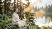 Young woman meditating by serene mountain lake at sunrise, practicing mindfulness in peaceful forest setting with golden sunlight filtering through trees.