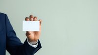 Businessman in navy suit holding blank white business card with hand visible against neutral gray background, ready for contact information or branding.