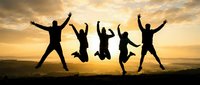 Group of five friends jumping with raised arms against golden sunset sky celebrating success and freedom outdoors on mountain landscape horizon.