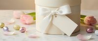 Elegant white gift box with satin ribbon and blank tag surrounded by pink tulips and colorful Easter eggs on soft neutral background for spring celebrations.