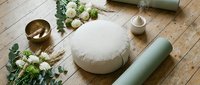 Flat lay yoga and meditation setup with white bolster pillow, sage green mat roll, brass bowl, white flowers, and greenery on wooden floor.