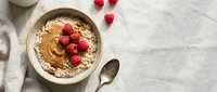 Oatmeal bowl topped with peanut butter, fresh raspberries, and banana slices on marble surface with spoon and cloth napkin for breakfast or healthy eating content.
