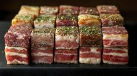 Raw marbled meat cubes seasoned with herbs and spices arranged in rows on dark stone surface, showcasing various cuts of premium beef and pork for gourmet cooking.
