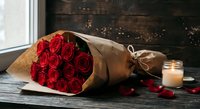 Red roses bouquet wrapped in brown paper with candle on wooden table near window creating romantic Valentine's Day or anniversary gift atmosphere.