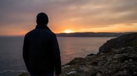Silhouetted figure in dark jacket standing on rocky coastal cliff overlooking ocean at golden sunset, contemplative mood for travel and lifestyle content.