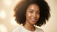 Young African American woman with curly hair smiling warmly at camera in soft natural lighting for beauty and lifestyle content.
