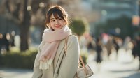 Young Asian woman in beige coat and pink scarf walking outdoors in urban setting with warm sunlight and blurred city background for lifestyle content.