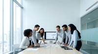 Diverse business team collaborating around conference table with computer displaying charts in modern office meeting room with city view.
