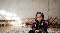 Young girl of Central Asian ethnicity wearing traditional embroidered clothing and braided hairstyle, sitting on ornate carpet in rustic interior with natural light. Cultural heritage portrait.