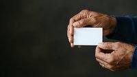Mature man with brown skin holding blank white business card in both hands against dark background for corporate branding.