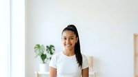Young woman with long dark hair and medium skin tone wearing white t-shirt, smiling at camera in bright minimalist interior with potted plant, suitable for lifestyle and wellness content.