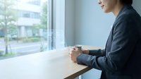 Professional businesswoman in dark suit holding coffee cup at modern office window overlooking urban cityscape during daytime work break.