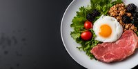 Healthy protein breakfast with fried egg, raw beef steak, quinoa, fresh berries, cherry tomatoes and green lettuce served on white plate, dark background.