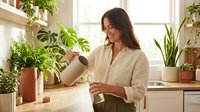 Young woman watering houseplants with spray bottle in bright kitchen surrounded by green foliage creating peaceful indoor gardening atmosphere.