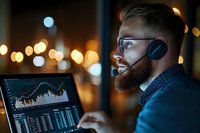 Young bearded Caucasian male trader in glasses and headphones analyzing financial market data on laptop at night with bokeh lights in background for stock trading concept.