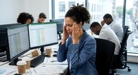 Stressed African American businesswoman experiencing headache while working on computer in busy corporate office environment with colleagues in background.