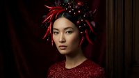 Young East Asian woman wearing red sequined sweater with elaborate red feathered headpiece adorned with sparkles against dark background for fashion and beauty editorial use.