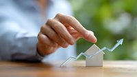 Caucasian businessman's hand pointing at upward trending arrow over miniature house model, representing real estate market growth and property investment success.