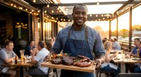 Smiling African American chef presenting grilled meat platter on outdoor restaurant terrace during golden hour with string lights and dining guests.