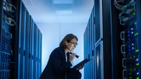 Professional Caucasian woman IT specialist with glasses working on tablet in modern server room with blue lighting and network equipment racks.
