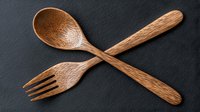 Crossed wooden spoon and fork utensils on dark background, handcrafted from natural oak wood with detailed grain texture, rustic kitchen decor elements.
