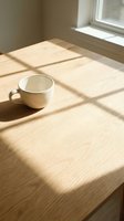 White ceramic mug on wooden table with natural sunlight streaming through window, creating geometric shadows in minimalist interior space.