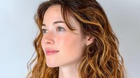 Young Caucasian woman with wavy brown hair highlighted with caramel balayage looking away thoughtfully against light background, natural makeup and glowing skin.
