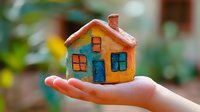 Colorful miniature clay house held in open palm against blurred green background, symbolizing home ownership and property concepts.