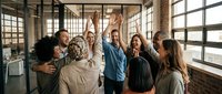 Diverse group of young professionals celebrating success with raised hands in modern loft office space with large windows, teamwork and achievement concept for corporate business projects.
